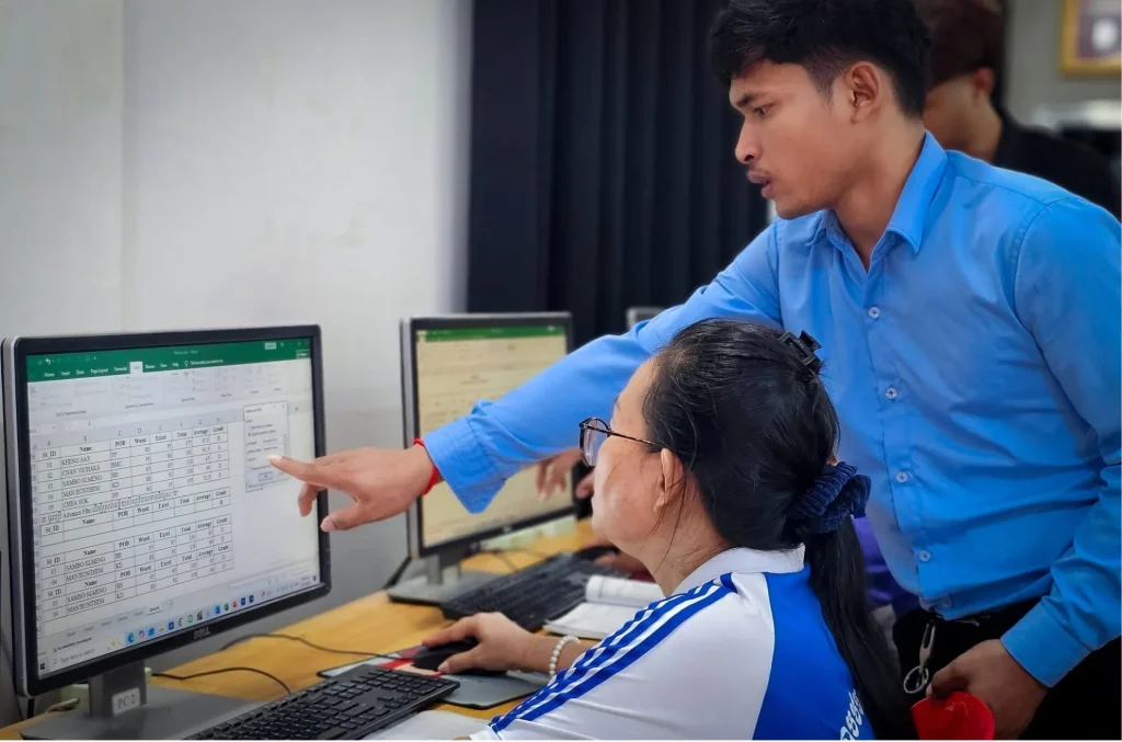 Chom Phanith pointing to a spreadsheet on a computer screen beside a seated learner.