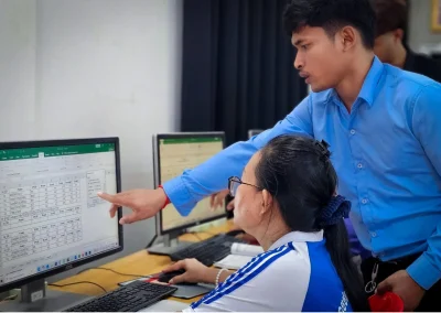 Chom Phanith pointing to a spreadsheet on a computer screen beside a seated learner.