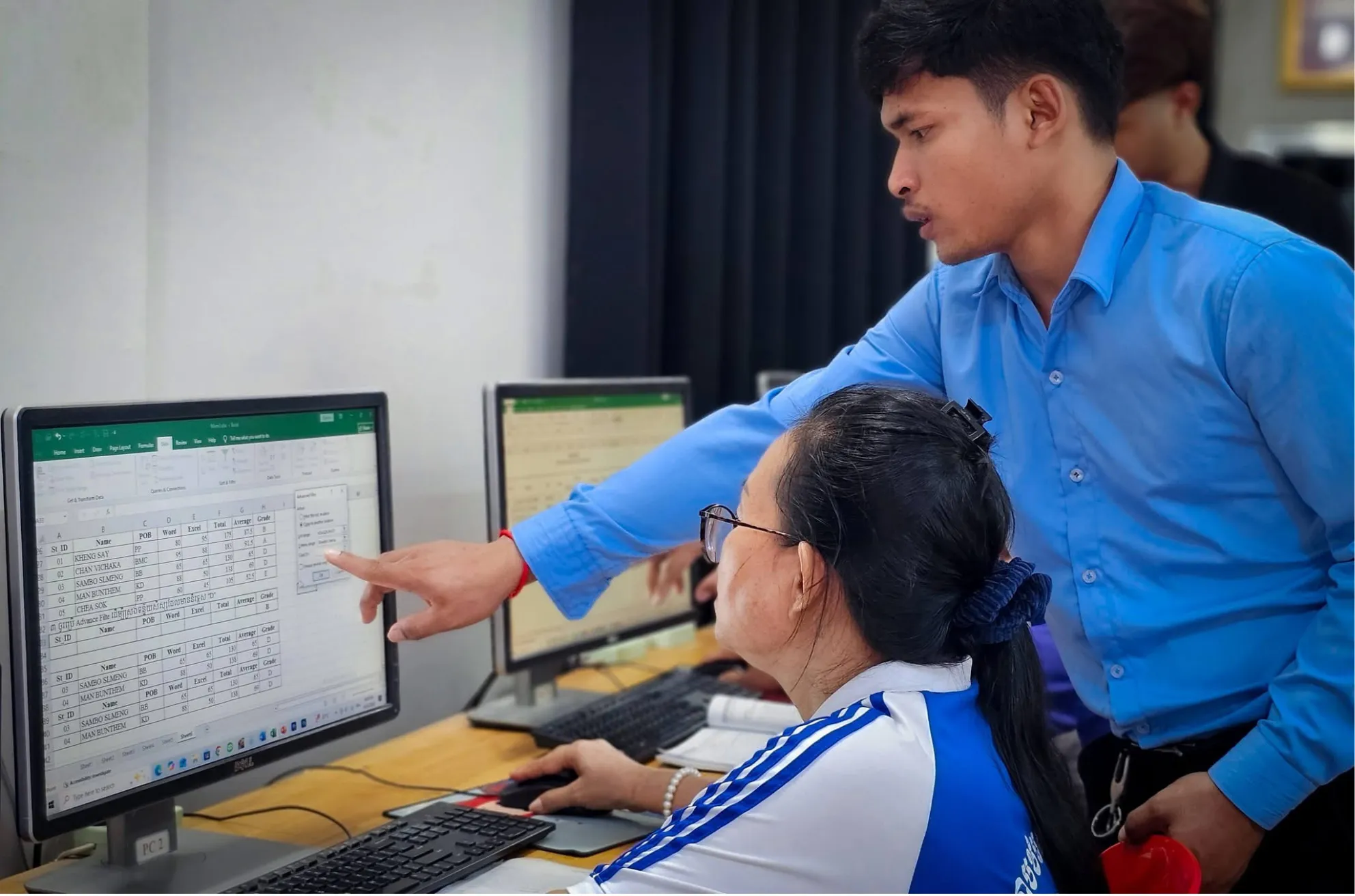 Chom Phanith pointing to a spreadsheet on a computer screen beside a seated learner.