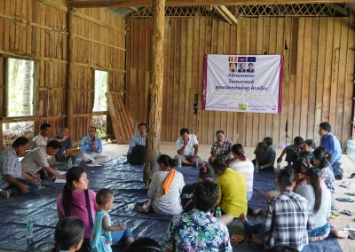 Community members sit in a circle on a floor mat during a discussion in a wooden meeting hall.