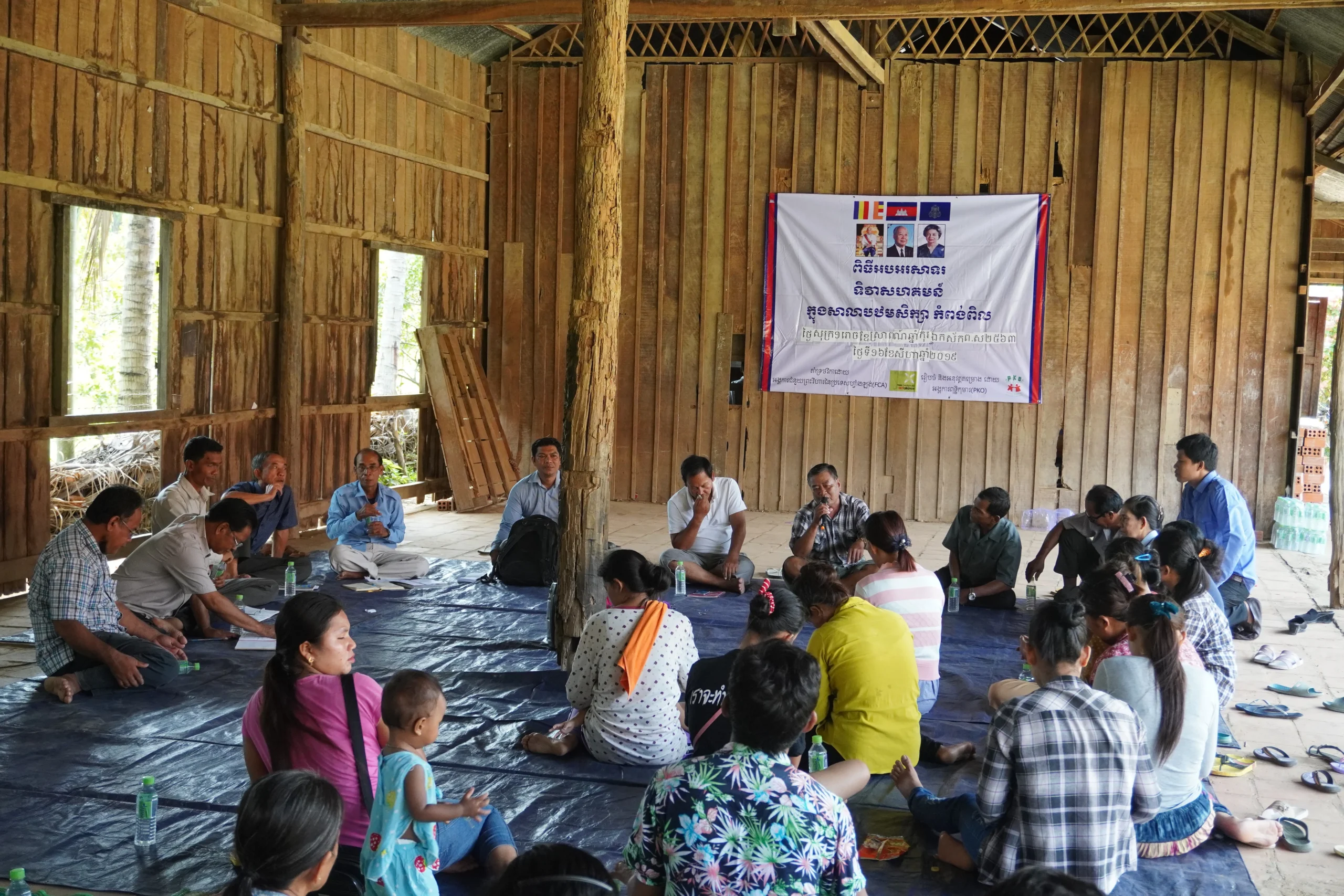 Community members sit in a circle on a floor mat during a discussion in a wooden meeting hall.