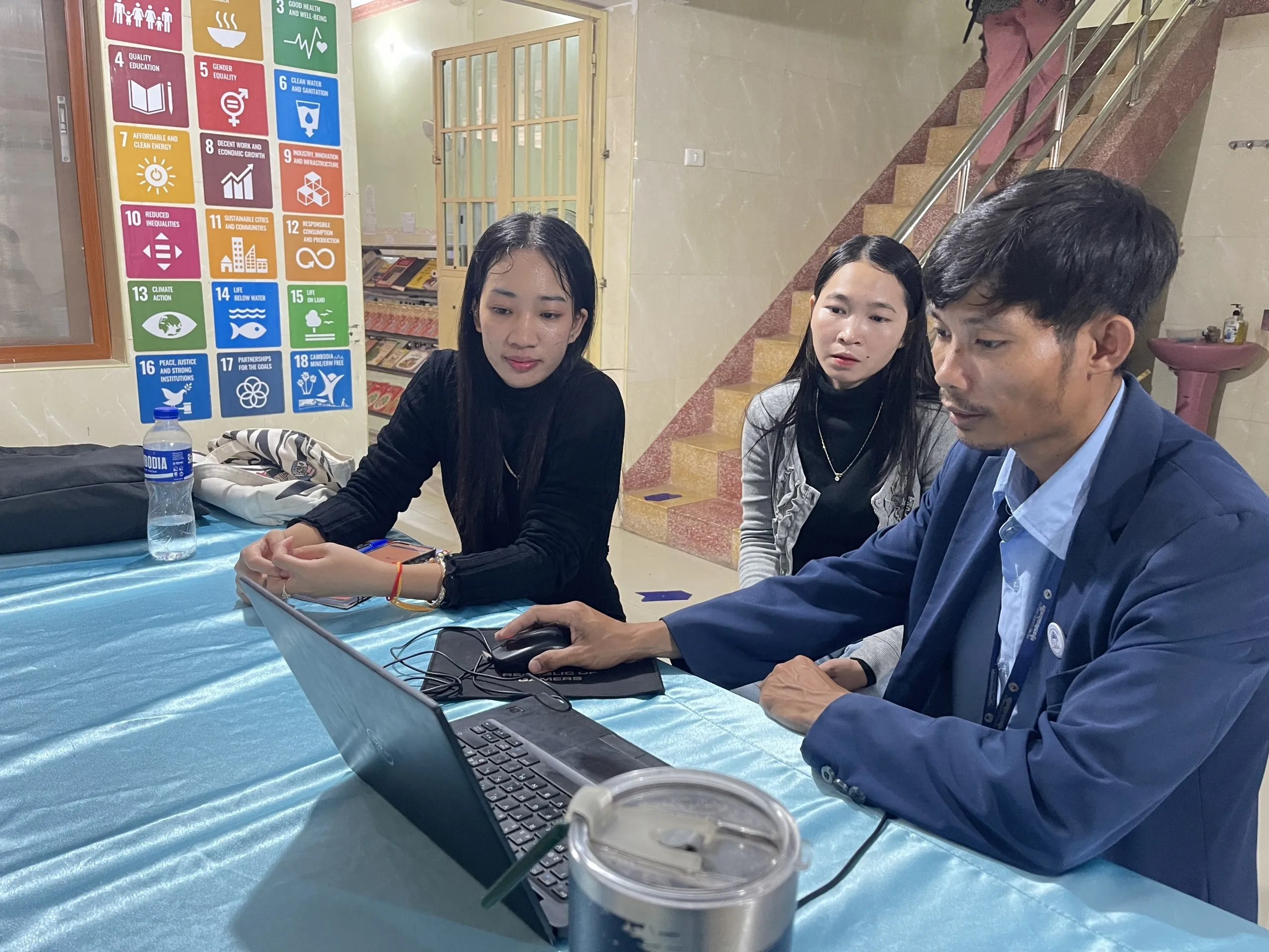 pko-consultation-meeting Three participants review a laptop together during a consultation meeting, with Sustainable Development Goals posters in the background.
